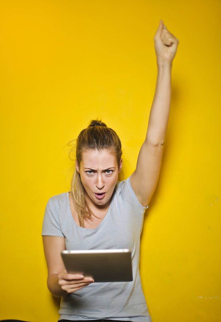 Joyful woman raises arm in celebration while holding a tablet against a vibrant yellow backdrop.